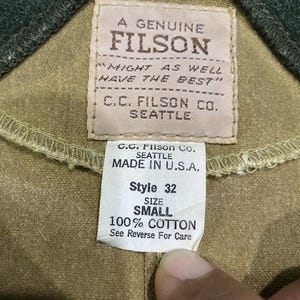 Peut inclure: Gros plan sur une &eacute;tiquette de v&ecirc;tement avec le texte "A GENUINE FILSON" et "C.C. FILSON CO. SEATTLE". L'&eacute;tiquette indique &eacute;galement "MADE IN U.S.A.", "Style 32", "SIZE SMALL" et "100% COTTON". Le v&ecirc;tement est de couleur beige.
