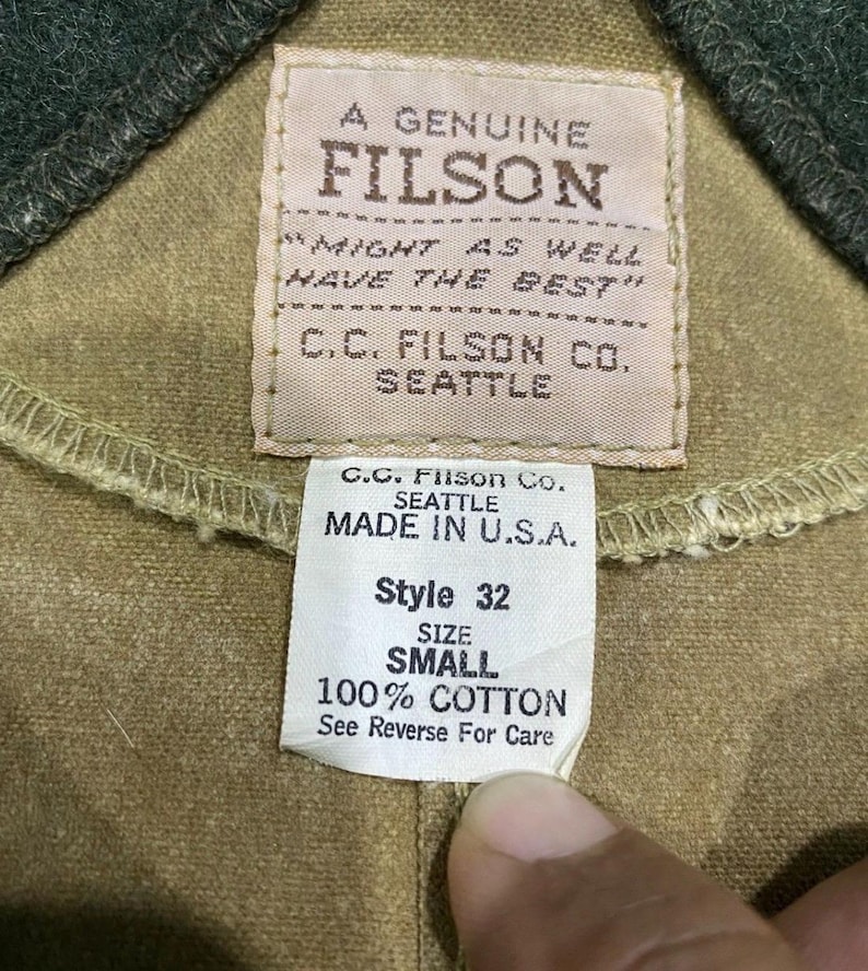 Peut inclure: Gros plan sur une &eacute;tiquette de v&ecirc;tement avec le texte "A GENUINE FILSON" et "C.C. FILSON CO. SEATTLE". L'&eacute;tiquette indique &eacute;galement "MADE IN U.S.A.", "Style 32", "SIZE SMALL" et "100% COTTON". Le v&ecirc;tement est de couleur beige.