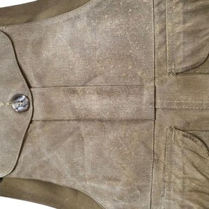 Peut inclure: Gros plan d'un gilet en toile cir&eacute;e de couleur vert olive. Le gilet est dot&eacute; d'une fermeture boutonn&eacute;e, de deux poches et d'un col vert fonc&eacute;. Le tissu a un aspect textur&eacute; et vieilli.