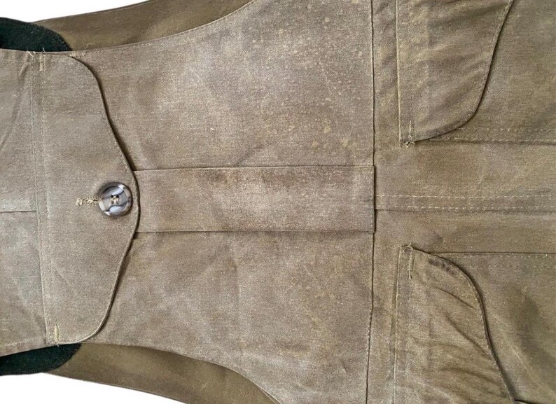 Peut inclure: Gros plan d'un gilet en toile cir&eacute;e de couleur vert olive. Le gilet est dot&eacute; d'une fermeture boutonn&eacute;e, de deux poches et d'un col vert fonc&eacute;. Le tissu a un aspect textur&eacute; et vieilli.