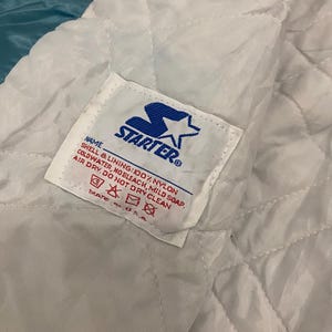 Puede incluir: Primer plano del interior de una chaqueta Starter vintage. El forro es blanco, acolchado y presenta una etiqueta con el logotipo de Starter e instrucciones de cuidado. El exterior de la chaqueta es de color turquesa.