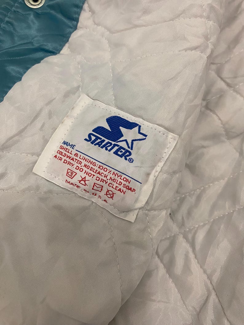 Puede incluir: Primer plano del interior de una chaqueta Starter vintage. El forro es blanco, acolchado y presenta una etiqueta con el logotipo de Starter e instrucciones de cuidado. El exterior de la chaqueta es de color turquesa.