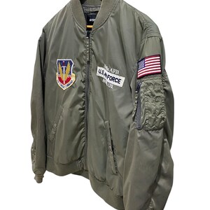 May include: An olive green bomber jacket featuring a front zipper, ribbed collar, and cuffs. The chest displays an Air Combat Command patch and a white rectangular patch reading Nellis AFB U.S. AIR FORCE AV. LTD. A small American flag patch is on the sleeve.