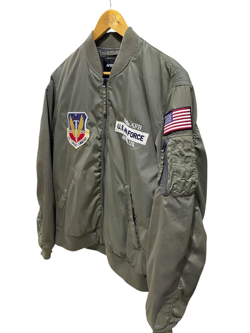 May include: An olive green bomber jacket featuring a front zipper, ribbed collar, and cuffs. The chest displays an Air Combat Command patch and a white rectangular patch reading Nellis AFB U.S. AIR FORCE AV. LTD. A small American flag patch is on the sleeve.