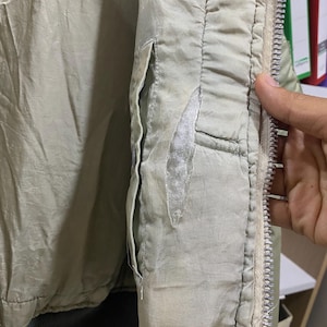 May include: Close-up of a light green jacket with a silver zipper and a pocket. The jacket shows signs of wear, with a tear in the fabric near the pocket. The interior lining is visible.