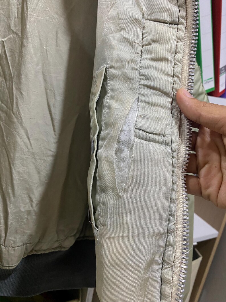 May include: Close-up of a light green jacket with a silver zipper and a pocket. The jacket shows signs of wear, with a tear in the fabric near the pocket. The interior lining is visible.