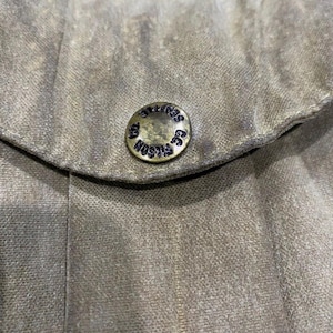 Peut inclure: Gros plan sur un tissu vert olive avec un bouton. Le bouton m&eacute;tallique circulaire porte l'inscription "FILSON CO. SEATTLE" en bleu fonc&eacute;. Le tissu pr&eacute;sente un aspect textur&eacute;.