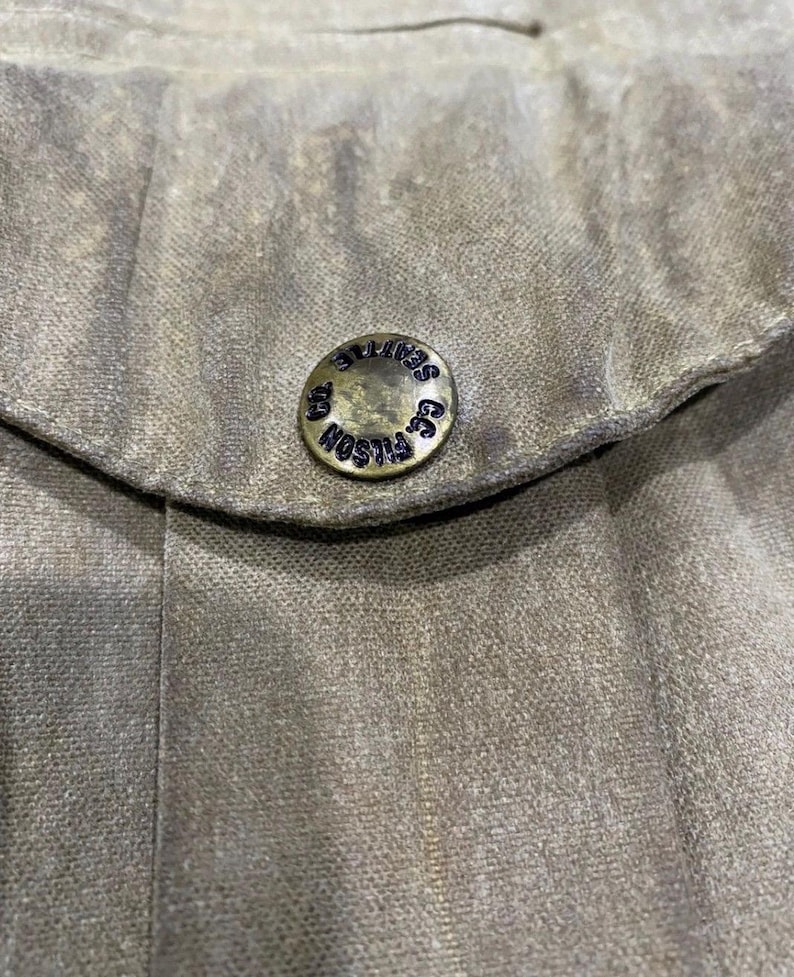 Peut inclure: Gros plan sur un tissu vert olive avec un bouton. Le bouton m&eacute;tallique circulaire porte l'inscription "FILSON CO. SEATTLE" en bleu fonc&eacute;. Le tissu pr&eacute;sente un aspect textur&eacute;.