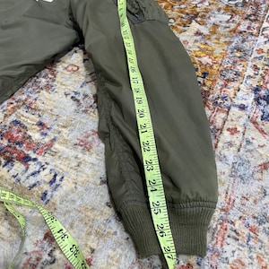 May include: An olive green bomber jacket with a U.S. Air Force Nellis AFB patch on the chest and an American flag patch on the sleeve. A yellow measuring tape is placed along the sleeve, showing a length of approximately 27 inches.