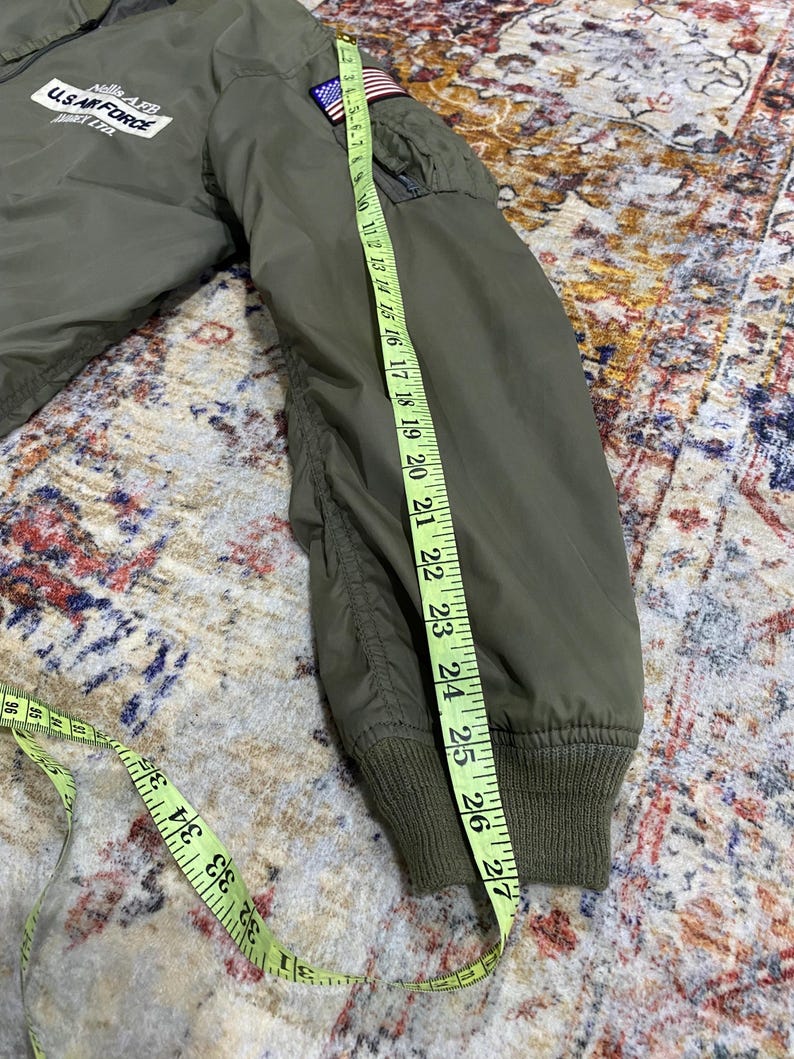 May include: An olive green bomber jacket with a U.S. Air Force Nellis AFB patch on the chest and an American flag patch on the sleeve. A yellow measuring tape is placed along the sleeve, showing a length of approximately 27 inches.