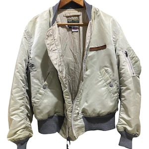 May include: A light beige bomber jacket with a gray collar, cuffs, and waistband. The jacket has a zipper closure, multiple pockets, and a small patch on the chest. It is hanging on a wooden hanger.