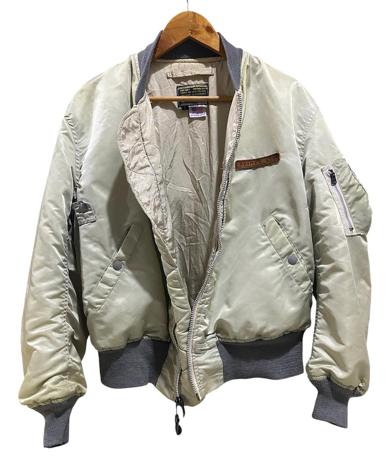 May include: A light beige bomber jacket with a gray collar, cuffs, and waistband. The jacket has a zipper closure, multiple pockets, and a small patch on the chest. It is hanging on a wooden hanger.