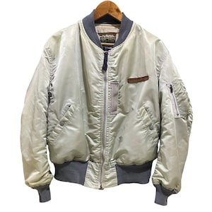 May include: A light green bomber jacket with a gray collar, cuffs, and waistband. The jacket has a zipper closure, two front pockets, and a small patch on the chest that reads "AVIREX". The jacket is hanging on a wooden hanger.