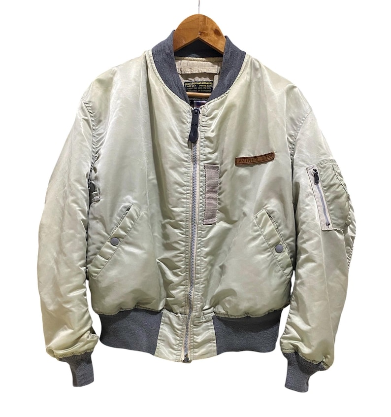 May include: A light green bomber jacket with a gray collar, cuffs, and waistband. The jacket has a zipper closure, two front pockets, and a small patch on the chest that reads "AVIREX". The jacket is hanging on a wooden hanger.