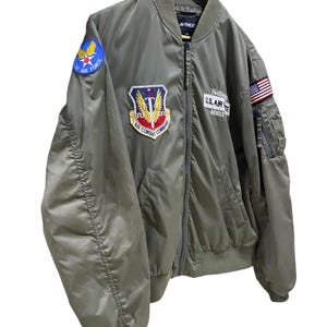 May include: An olive green bomber jacket featuring a front zipper, ribbed collar, cuffs, and hem. Patches include a blue Air Force emblem on the shoulder, an Air Combat Command patch on the chest, a Nellis U.S. Air Force Avirex patch, and a U.S. flag.