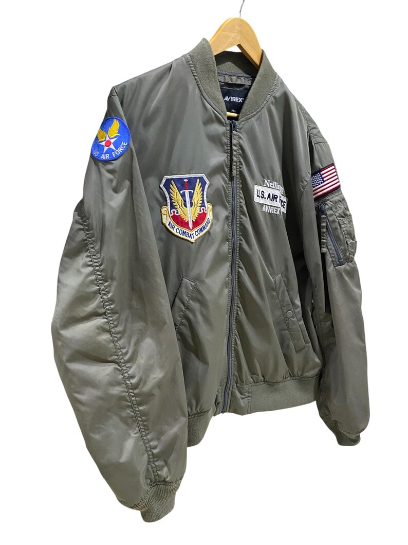 May include: An olive green bomber jacket featuring a front zipper, ribbed collar, cuffs, and hem. Patches include a blue Air Force emblem on the shoulder, an Air Combat Command patch on the chest, a Nellis U.S. Air Force Avirex patch, and a U.S. flag.