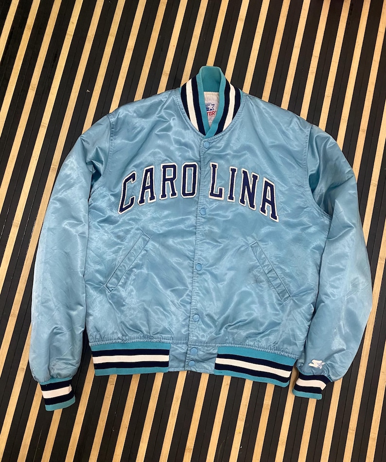 Puede incluir: Chaqueta de sat&eacute;n azul claro con la palabra "CAROLINA" en letras azules marino en el pecho. La chaqueta tiene cuello, pu&ntilde;os y dobladillo acanalados con rayas azul marino y blancas. Un logotipo de Starter es visible en el pu&ntilde;o.