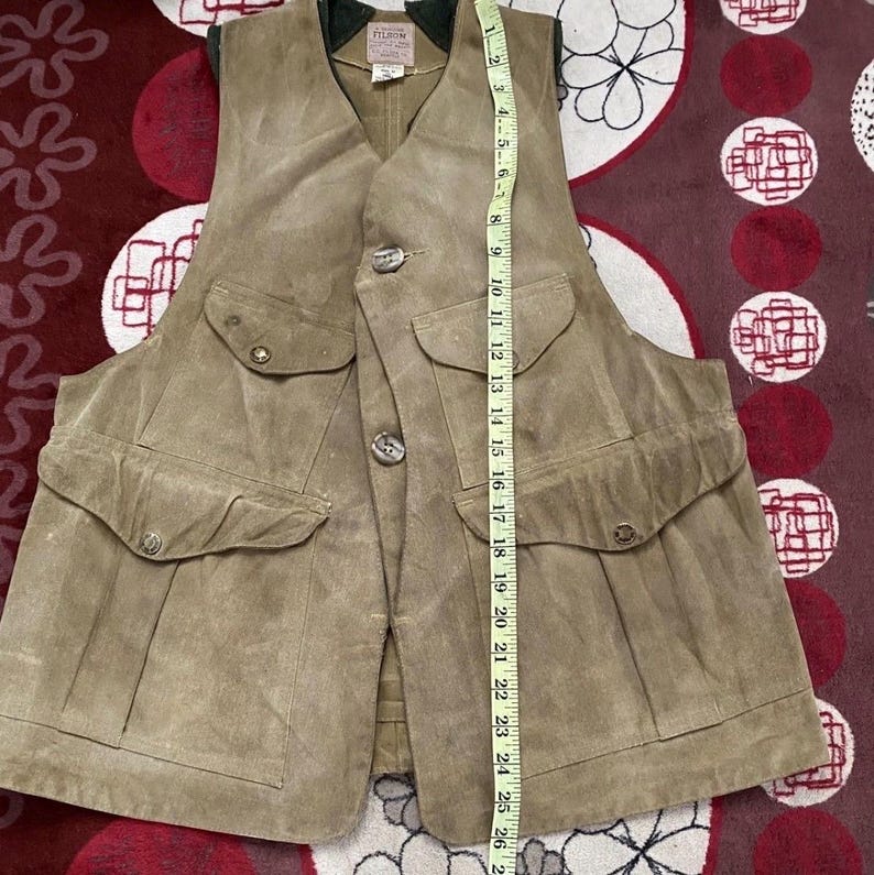 Peut inclure: Un gilet en daim beige avec quatre poches avant et des fermetures boutonn&eacute;es. Le gilet a un col en V et un col vert. Un m&egrave;tre ruban est plac&eacute; &agrave; c&ocirc;t&eacute; du gilet, indiquant une longueur d'environ 66 cm.