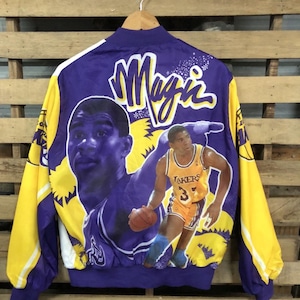 Puede incluir: Una chaqueta reversible de color morado y amarillo con una gran imagen de Magic Johnson vistiendo una camiseta de los Los Angeles Lakers. La chaqueta tiene la palabra "Magic" escrita en grandes letras blancas en la parte posterior.