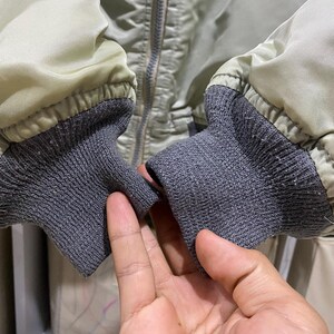 May include: Close-up of a light green bomber jacket with gray ribbed cuffs. The jacket has a silver zipper and a quilted texture. The cuffs are made of a gray, textured knit fabric.