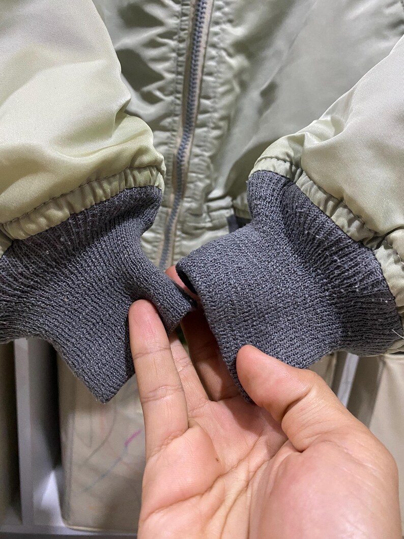 May include: Close-up of a light green bomber jacket with gray ribbed cuffs. The jacket has a silver zipper and a quilted texture. The cuffs are made of a gray, textured knit fabric.