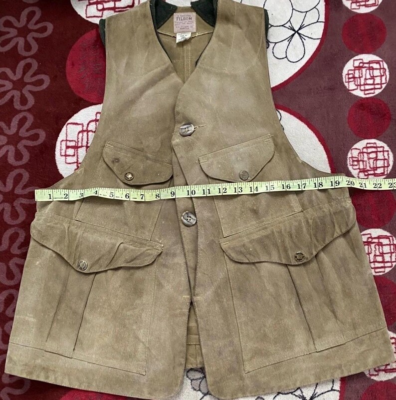 Peut inclure: Un gilet en daim beige avec quatre poches avant et des fermetures boutonn&eacute;es. Le gilet a un col vert fonc&eacute; et une &eacute;tiquette qui dit "FILSON". Le gilet mesure environ 51 cm de large au niveau de la poitrine.