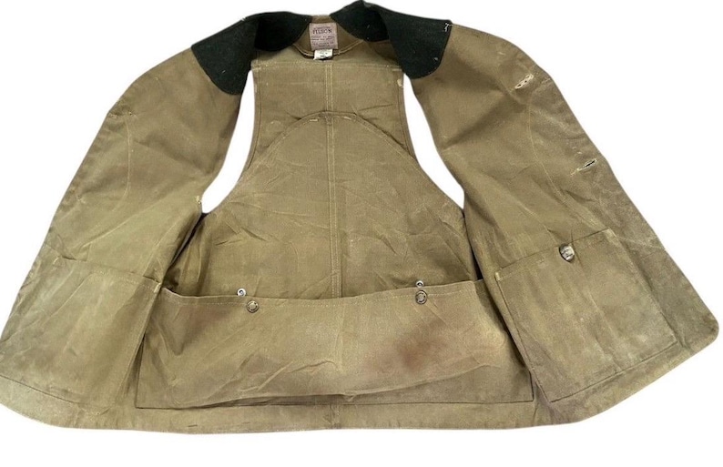 Peut inclure: Un gilet ouvert de couleur beige avec plusieurs poches et un col noir. Le gilet a un aspect us&eacute;, avec des coutures visibles et des fermetures &agrave; boutons m&eacute;talliques. Le design sugg&egrave;re un th&egrave;me de v&ecirc;tements de travail ou de plein air.