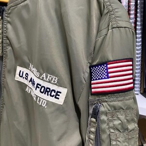 May include: An olive green bomber jacket featuring a white rectangular patch with the text Nellis AFB, U.S. AIR FORCE, and AVIREX LTD. in dark blue. A rectangular embroidered American flag patch is on the sleeve above a zippered utility pocket.