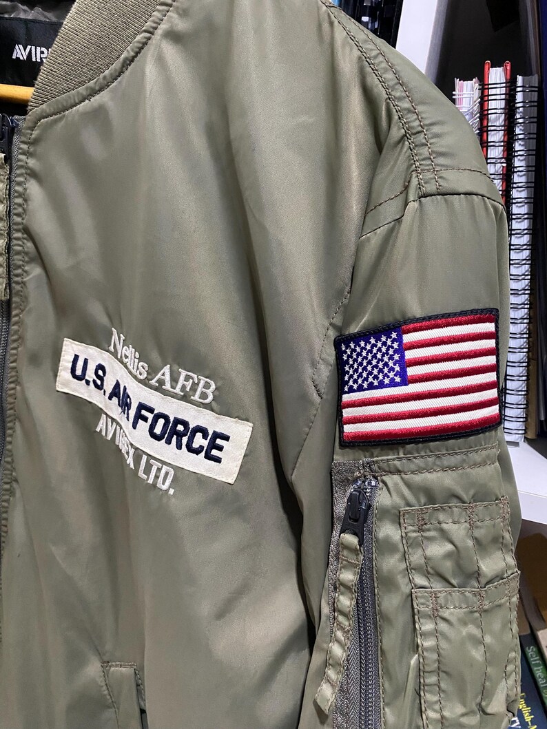 May include: An olive green bomber jacket featuring a white rectangular patch with the text Nellis AFB, U.S. AIR FORCE, and AVIREX LTD. in dark blue. A rectangular embroidered American flag patch is on the sleeve above a zippered utility pocket.