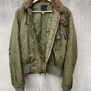 May include: Olive green bomber jacket with a brown faux fur collar, a quilted interior, and ribbed cuffs and hem. The jacket has a zipper closure and two front pockets. It is hanging on a wooden hanger against a gray concrete wall.