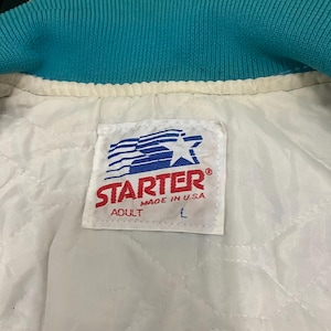 Puede incluir: Primer plano del interior de una chaqueta Starter turquesa y blanca. El cuello de la chaqueta es turquesa y el forro es blanco con un patr&oacute;n acolchado. Una etiqueta Starter es visible, con el texto "STARTER MADE IN U.S.A. ADULT L".