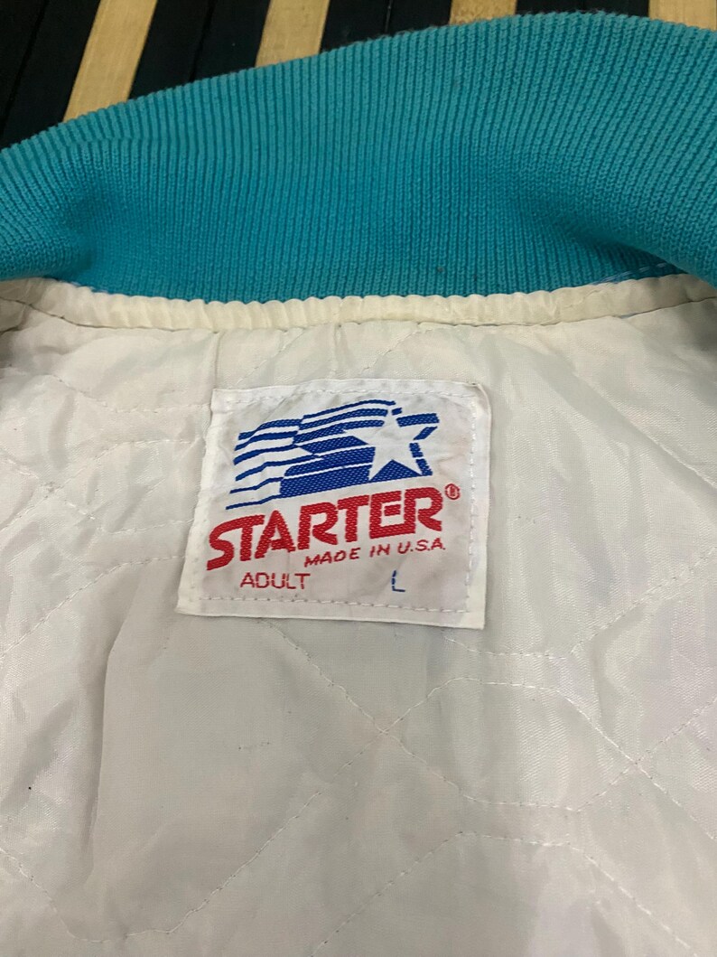 Puede incluir: Primer plano del interior de una chaqueta Starter turquesa y blanca. El cuello de la chaqueta es turquesa y el forro es blanco con un patr&oacute;n acolchado. Una etiqueta Starter es visible, con el texto "STARTER MADE IN U.S.A. ADULT L".
