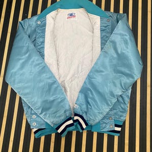 Puede incluir: Chaqueta de sat&eacute;n azul claro con cuello y pu&ntilde;os turquesa. La chaqueta tiene un forro acolchado blanco y una etiqueta de la marca Starter. Los pu&ntilde;os presentan rayas azul marino y blancas. La chaqueta es abotonada y tiene un peque&ntilde;o logotipo de estrella en la manga.