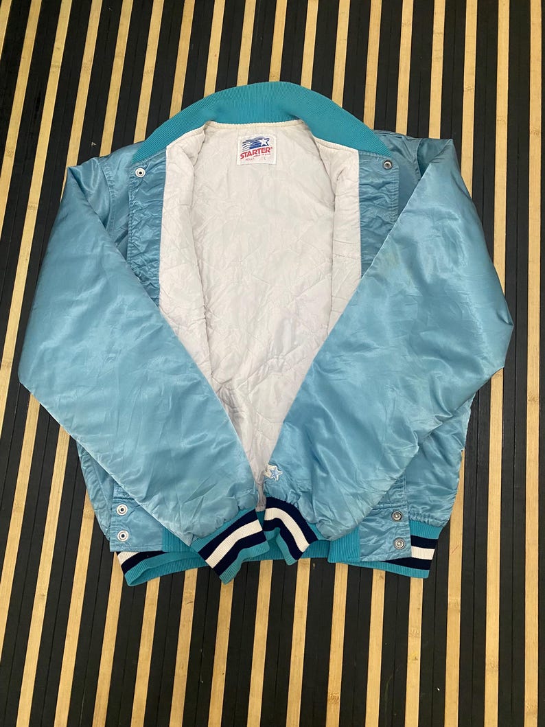Puede incluir: Chaqueta de sat&eacute;n azul claro con cuello y pu&ntilde;os turquesa. La chaqueta tiene un forro acolchado blanco y una etiqueta de la marca Starter. Los pu&ntilde;os presentan rayas azul marino y blancas. La chaqueta es abotonada y tiene un peque&ntilde;o logotipo de estrella en la manga.