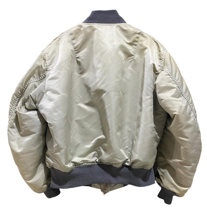 May include: Back view of a light green bomber jacket with a gray ribbed collar, cuffs, and hem. The jacket has a shiny, satin-like appearance and a classic bomber silhouette. The jacket is a neutral color.
