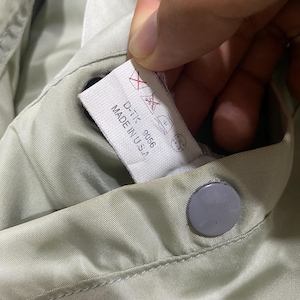 May include: Close-up of a light green garment with a tag that reads "D-TK 9056 MADE IN U.S.A." The tag also features care symbols. A gray button is visible.