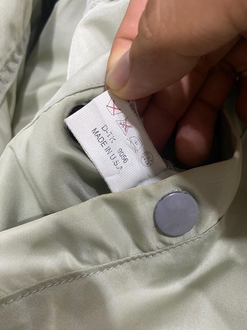 May include: Close-up of a light green garment with a tag that reads "D-TK 9056 MADE IN U.S.A." The tag also features care symbols. A gray button is visible.