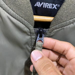 May include: An olive green bomber jacket featuring a ribbed collar and a black interior label with the white text AVIREX. The jacket includes a black metal zipper pull engraved with the brand name AVIREX and a matching olive green fabric pull tab.