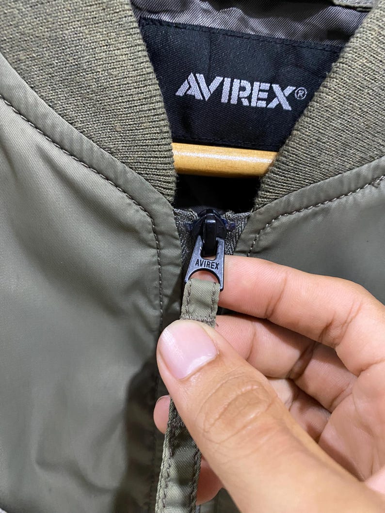 May include: An olive green bomber jacket featuring a ribbed collar and a black interior label with the white text AVIREX. The jacket includes a black metal zipper pull engraved with the brand name AVIREX and a matching olive green fabric pull tab.