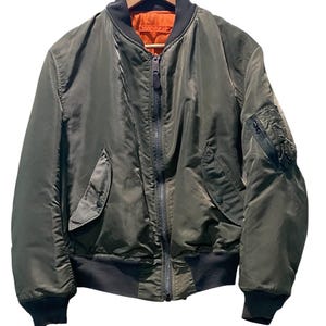Puede incluir: Una chaqueta tipo bomber de color verde oliva con una cremallera central de metal, cuello, puños y dobladillo de canalé negro. La prenda incluye dos bolsillos delanteros inclinados con cierre de corchete y un bolsillo utilitario con cremallera en la manga izquierda, con un forro de color naranja brillante.