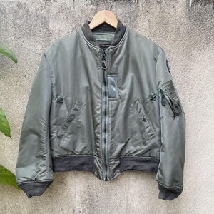 May include: A sage green bomber jacket with a dark grey collar, cuffs, and waistband. The jacket has a zip closure, multiple pockets, and a small patch on the sleeve. The jacket is hanging on a wooden hanger.