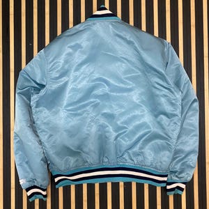 Puede incluir: Chaqueta bomber de sat&eacute;n azul claro con cuello, pu&ntilde;os y dobladillo de canal&eacute;. La chaqueta presenta rayas blancas y azul marino en el cuello, los pu&ntilde;os y el dobladillo. Se muestra sobre un fondo de madera a rayas.