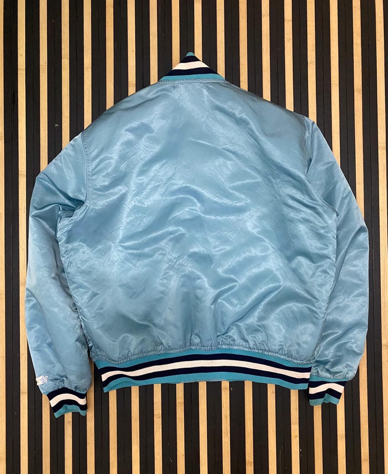 Puede incluir: Chaqueta bomber de sat&eacute;n azul claro con cuello, pu&ntilde;os y dobladillo de canal&eacute;. La chaqueta presenta rayas blancas y azul marino en el cuello, los pu&ntilde;os y el dobladillo. Se muestra sobre un fondo de madera a rayas.