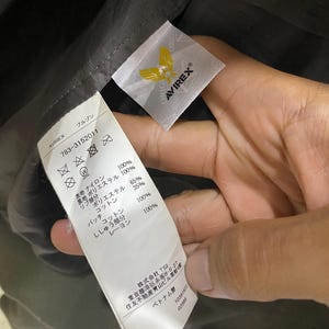 May include: A white fabric garment care label with black text and an Avirex logo featuring a yellow eagle. The label lists material compositions including nylon, polyester, cotton, and rayon, along with care symbols and manufacturing details.