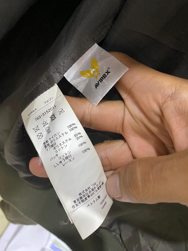 May include: A white fabric garment care label with black text and an Avirex logo featuring a yellow eagle. The label lists material compositions including nylon, polyester, cotton, and rayon, along with care symbols and manufacturing details.