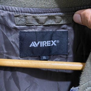 May include: An olive green jacket interior featuring a black rectangular brand label with the white text AVIREX and a smaller tag below it indicating size XL. The garment has a ribbed olive green collar and a smooth, dark brown lining fabric.
