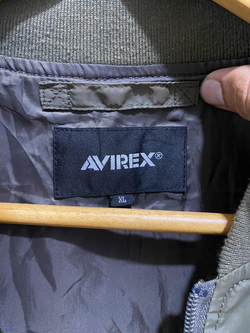 May include: An olive green jacket interior featuring a black rectangular brand label with the white text AVIREX and a smaller tag below it indicating size XL. The garment has a ribbed olive green collar and a smooth, dark brown lining fabric.