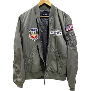 May include: An olive green bomber jacket featuring an Air Combat Command patch on the right chest and a U.S. Air Force Nellis AFB patch on the left chest. The left sleeve displays an American flag patch and a zippered utility pocket.