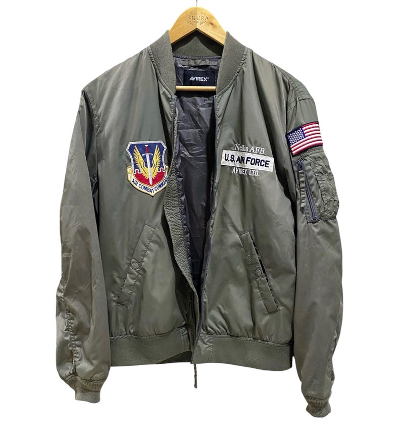 May include: An olive green bomber jacket featuring an Air Combat Command patch on the right chest and a U.S. Air Force Nellis AFB patch on the left chest. The left sleeve displays an American flag patch and a zippered utility pocket.