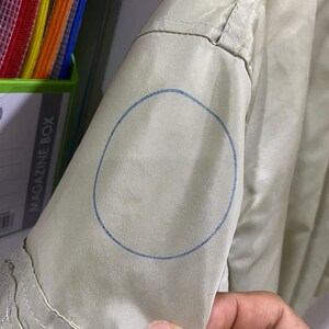 May include: Close-up of a light green jacket sleeve. A blue circle is drawn on the sleeve. The jacket has a pocket and a seam detail. The sleeve is being held by a person's hand.